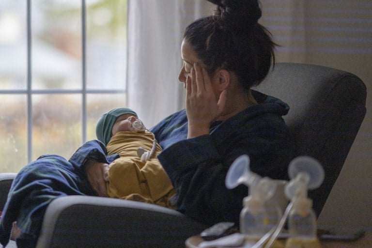A woman sits in an armchair near a window, holding a sleeping baby dressed in a yellow outfit and green hat. She rests one hand on her face. In the foreground, breast pump equipment is visible on a table alongside a book of postpartum affirmations. The room appears calm and quiet.