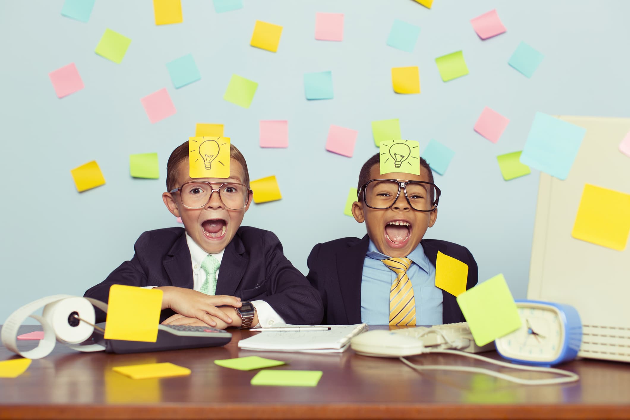 Two young businessmen sit at their office computer desk with ecstatic expressions on their faces and light bulb sticky notes on their foreheads. They are excited as they have thought of new business ideas. They are wearing a suit and tie and glasses.