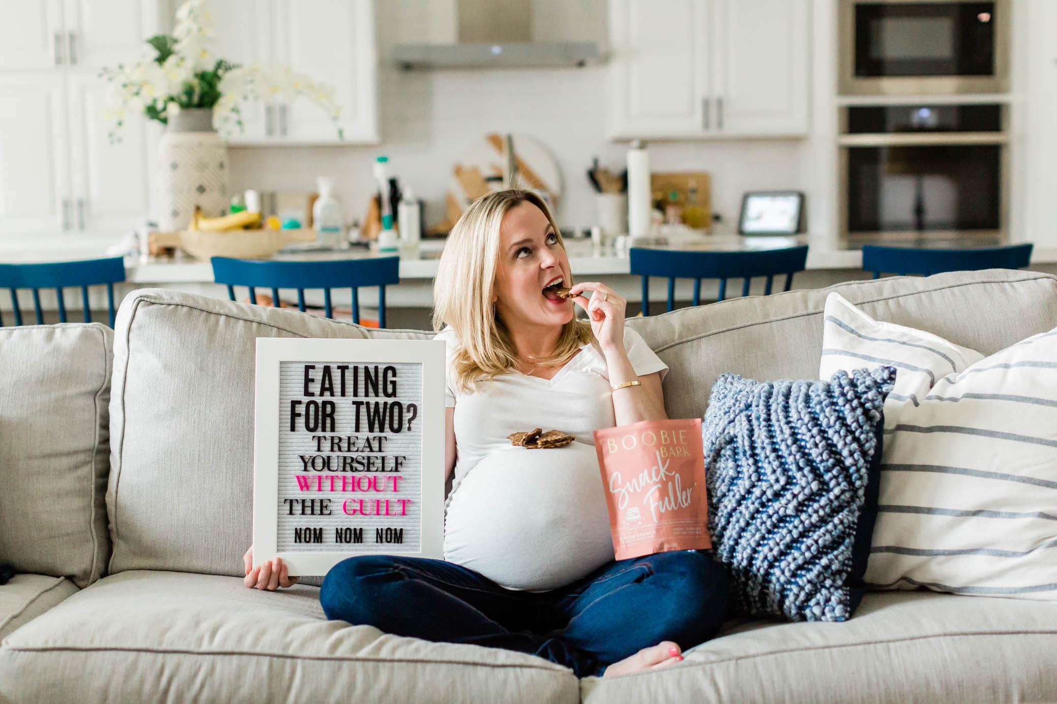 Pregnant woman sitting on a couch next to a letterboard eating Boobie Bark.