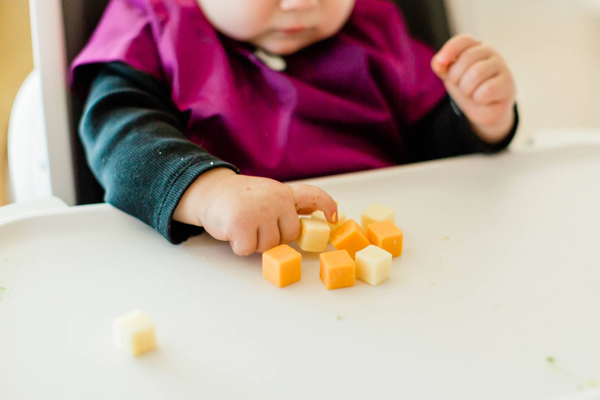 Cropped picture of a baby girl sitting in her high chair grabbing a cheese cube.