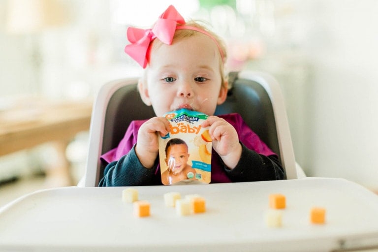 Baby girl eating a Stonyfield Yobaby pouch while sitting in her highchair with cubes of cheese on her tray.