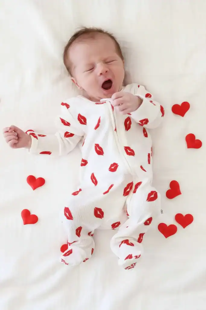 Baby yawning in a white onesie with red lips print, surrounded by small red hearts.