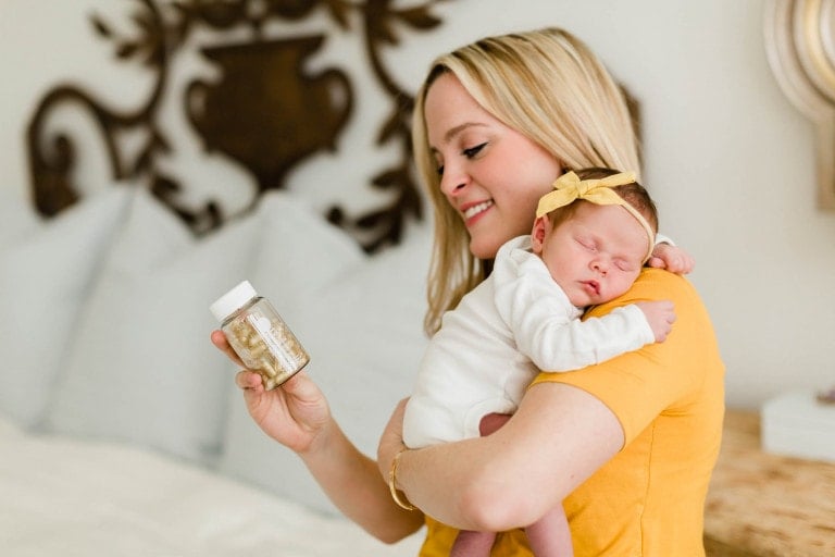 A woman with blonde hair, wearing a yellow top, smiles while holding a sleeping baby with a yellow bow on its head in one arm. In her other hand, she holds a bottle of postnatal multivitamins. They are in a room with a bed and decorative wall art in the background.