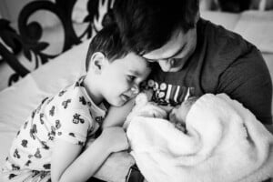 A man holds a newborn wrapped in a blanket while an older child in pajamas leans in close, smiling gently.