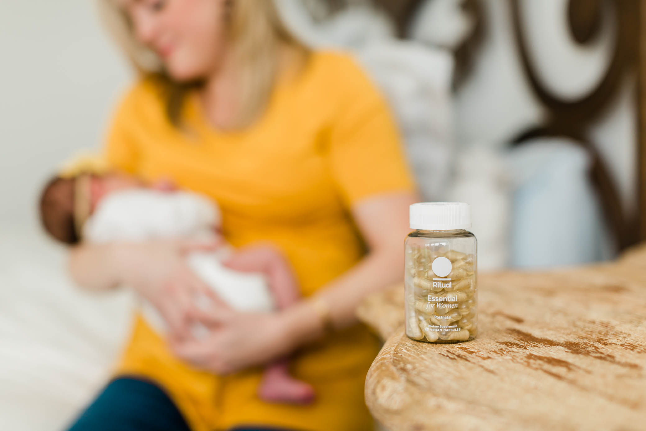 Ritual Postnatal multivitamin bottle with a mom breastfeeding her baby in the background.