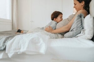 A young child rests their head on a pregnant persons belly while they both relax on a bed in a bright bedroom.