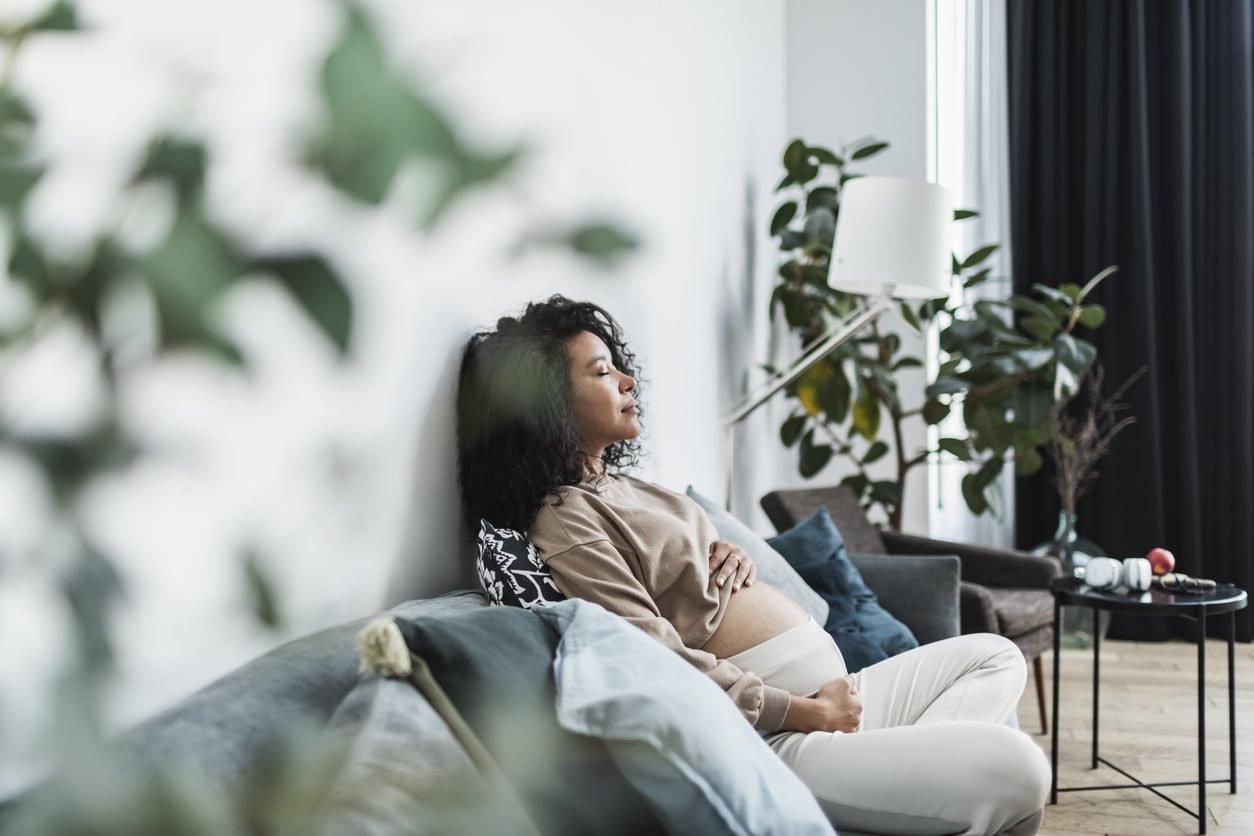 Pregnant black woman sitting on sofa with closed eyes, touching her belly and smile