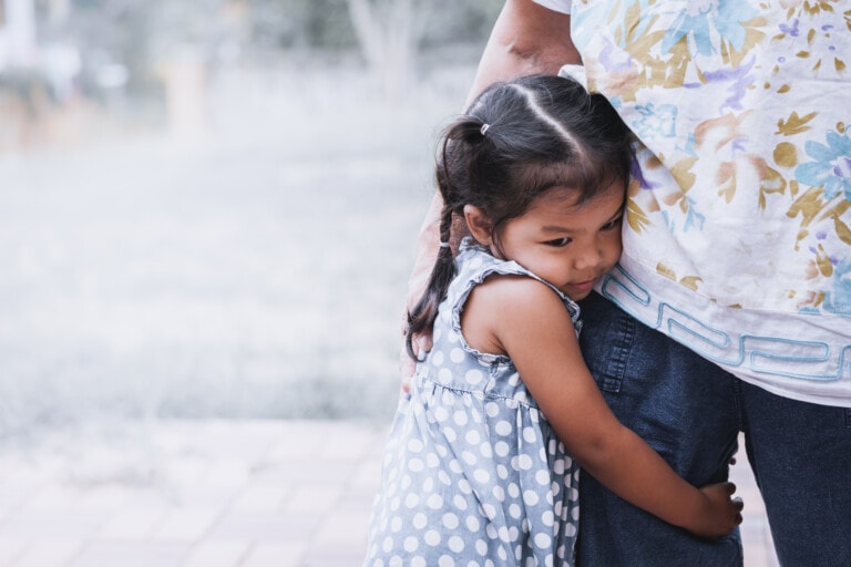 A young child with pigtails and a polka dot dress clings to an adult's leg, displaying signs of separation anxiety, while outdoors. The adult is wearing jeans and a floral-patterned shirt. The background is blurry, making the focus on the child and adult.