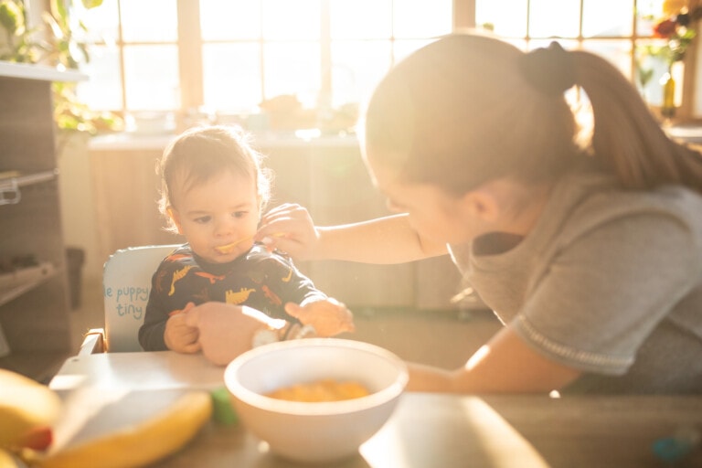 Teenage girl feeding baby boy with solid food indoors,baby sitting in a high chair