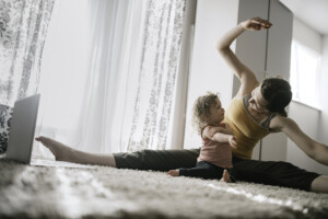 A mother does a virtual exercise class with her toddler aged daughter in their living room. The little girl copies her moms exercise routine. Part of the regular routine or the new normal with social distancing and Covid-19.