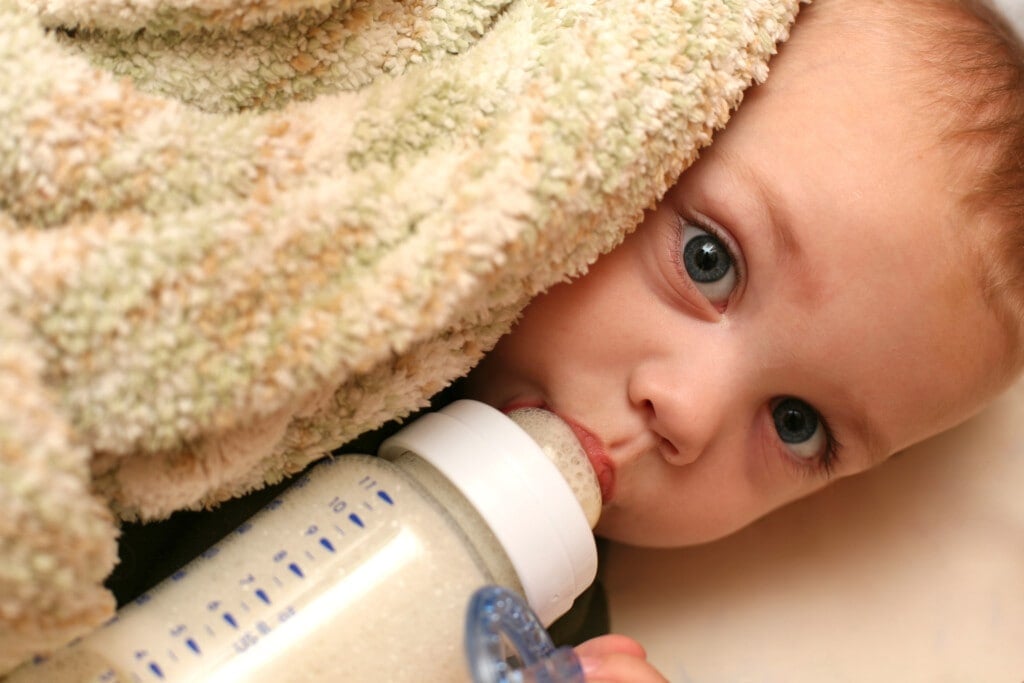 Close up of a blue eyed baby boy drinking his bottle of milk and cuddling with his blanket.