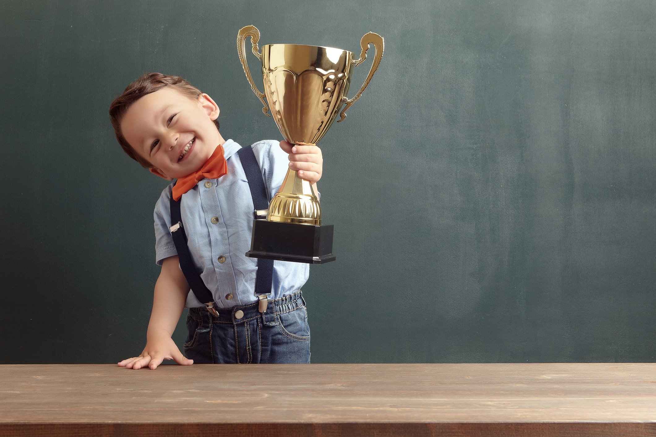 A cute, smiling 2-3 years old boy is standing behind a wooden table and raising a golden trophy with his hand. Little boy is wearing an orange bow tie and blue trousers with suspenders.
