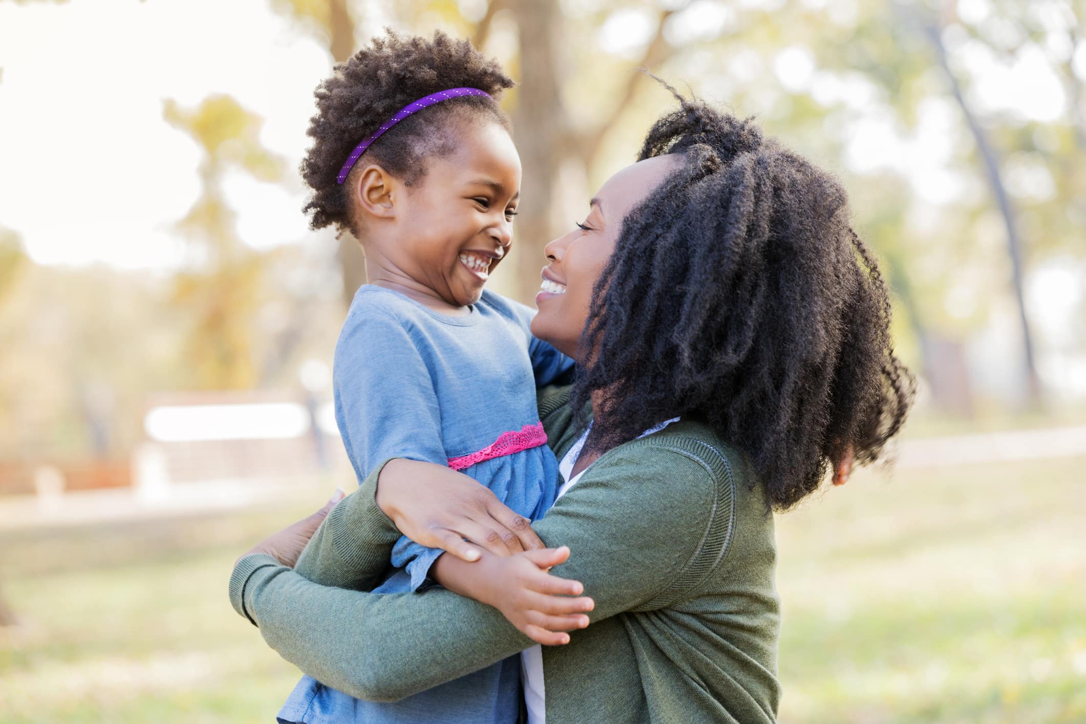 A mid adult mother stands in a sunny field outdoors and holds her daughter up. They smile at each other as they embrace.