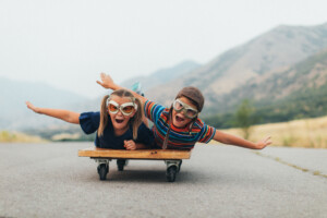 Two young children, a boy and a girl, are imagining flying into the sky while riding on a press cart. They have their arms spread out like wings and ready to use imagination in being like an airplane and piloting the airplane into the sky. They are wearing flying goggles on a rural road in Utah, USA.