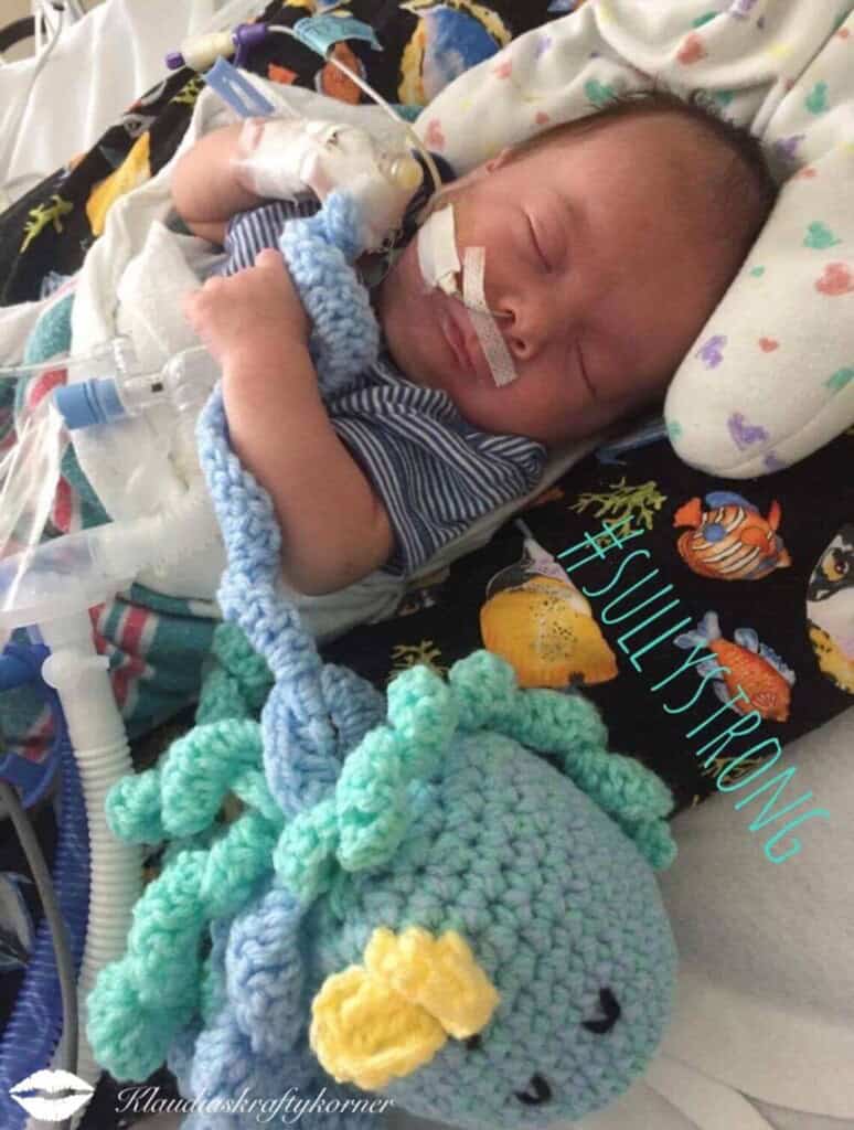 A sleeping baby with medical tubes is wearing a striped shirt and wrapped in a blanket adorned with sea creatures, holding a crocheted octopus toy. This tender scene, marked by #SullyStrong, captures the essence of thoughtful NICU gifts that bring comfort and hope.
