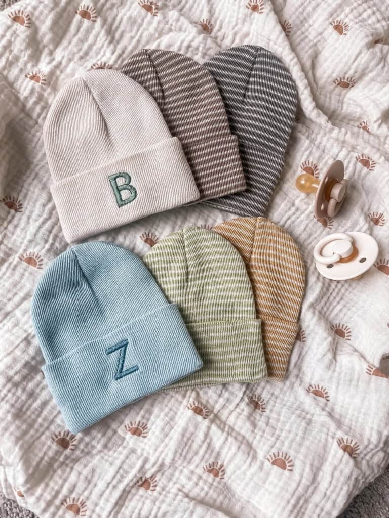 Five knitted baby hats, perfect as NICU gifts, rest on a patterned blanket. The hats, in shades of beige, brown, gray, blue, and green, feature embroidered letters B and Z. Nearby, two pacifiers add to the delightful arrangement.