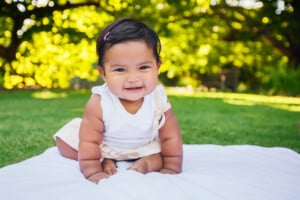 A baby wearing a white shirt and light-colored shorts is smiling while sitting on a white blanket on grass. The background is filled with green trees and bright sunlight. The baby, who appears to be the perfect example for those wondering "when do babies start sitting up," seems to be in a park or garden.