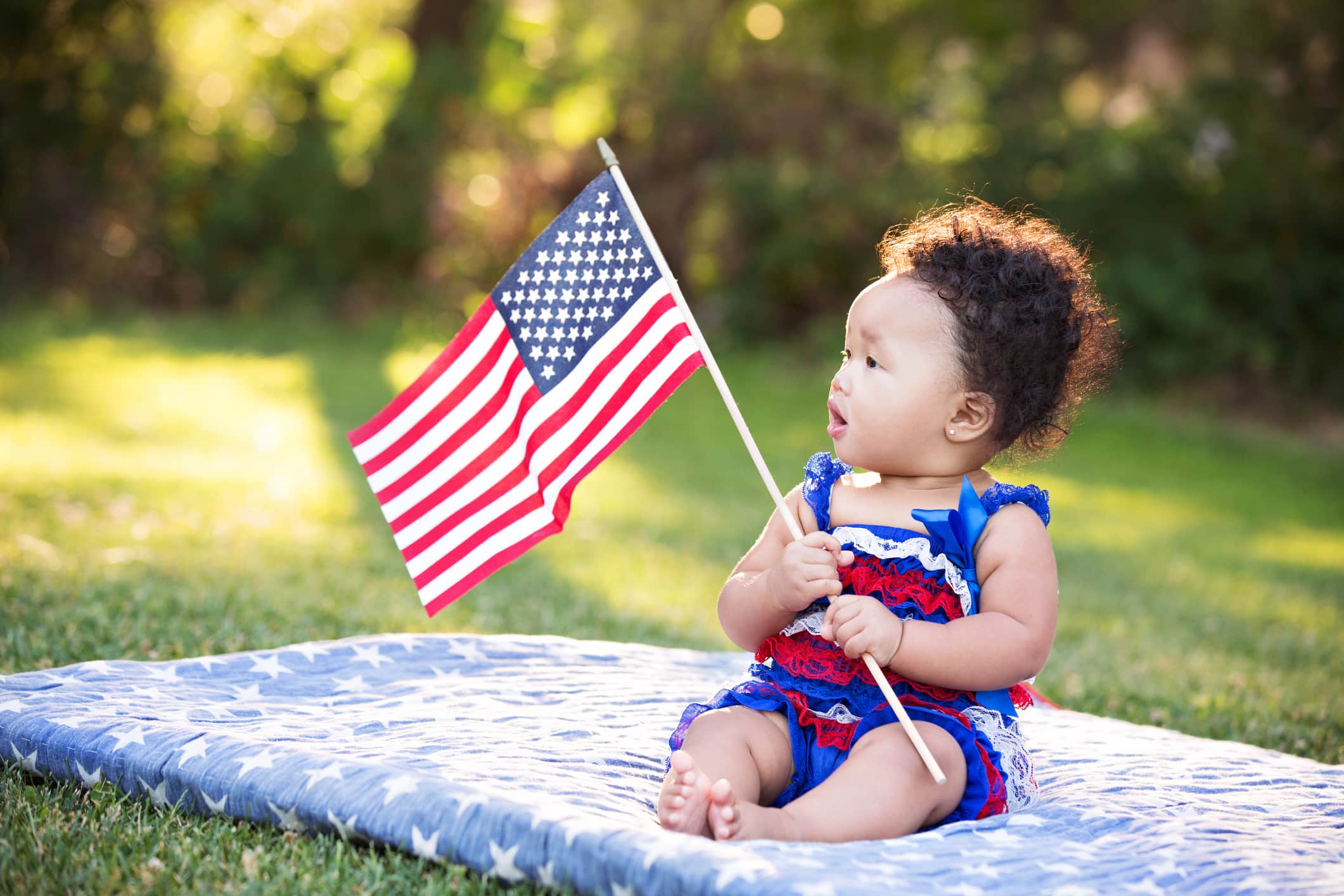 Beautiful six month old multi ethnic baby girl sitting on a blanket at the park and looking at the American flag she is holding.