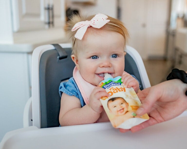 A baby with a light pink bow in her hair sits in a high chair, holding a pouch of baby food. An adult hand is helping her with the pouch. The baby is wearing a blue outfit and a pink bib. The background shows a kitchen setting, hinting that maybe this sweet little one needs yogurt next time.