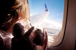 Baby girl sitting next to the airplane window looking out and holding her teddybear.
