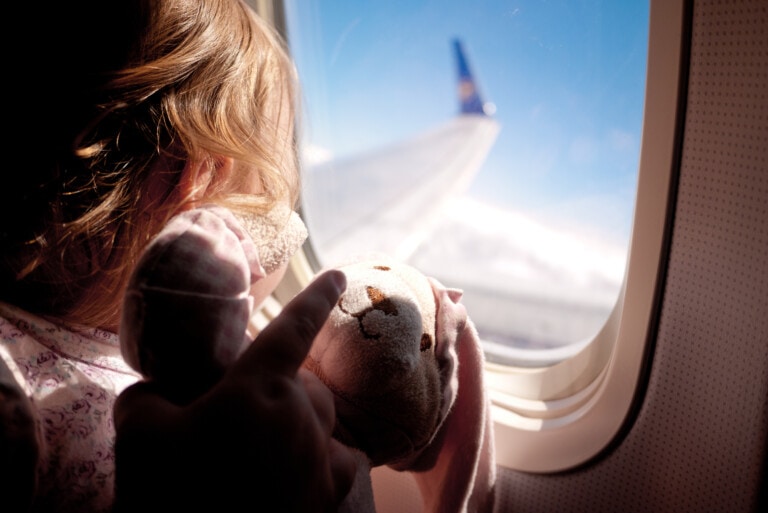 Baby girl sitting next to the airplane window looking out and holding her teddybear.