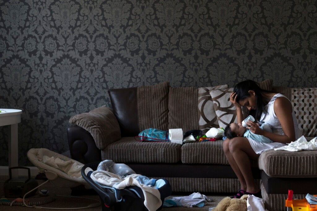 Stressed young mother sitting on her sofa whilst feeding her baby son. She has her head in her hand and is surrounded by mess.
