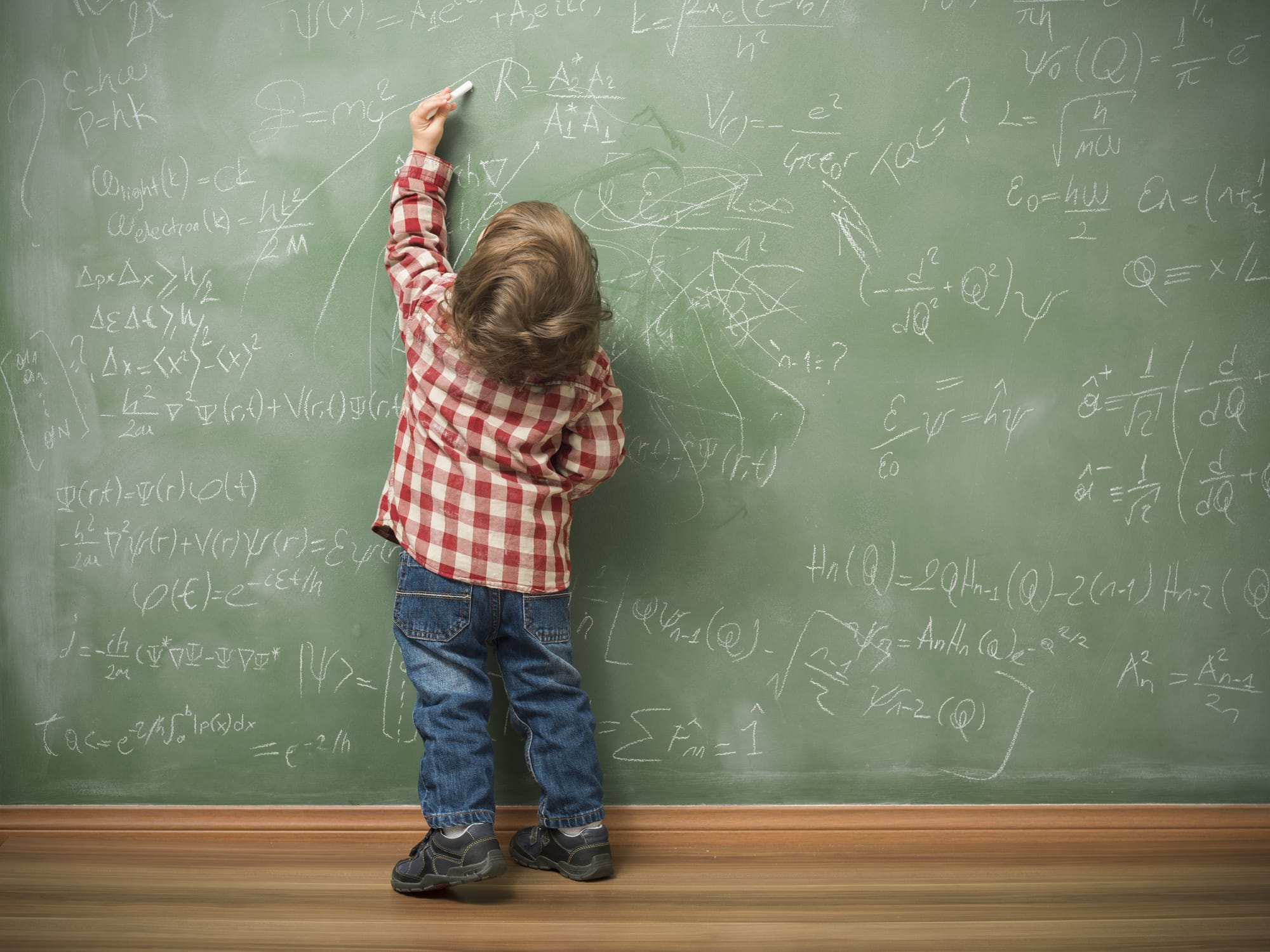 Little boy writing on green blackboard with math formulas written on. He is wearing a red plaid shirt and standing on the left side of frame. The green board is full of mathematics and physics formulas. He is seen in full length and holding chalk in left hand.