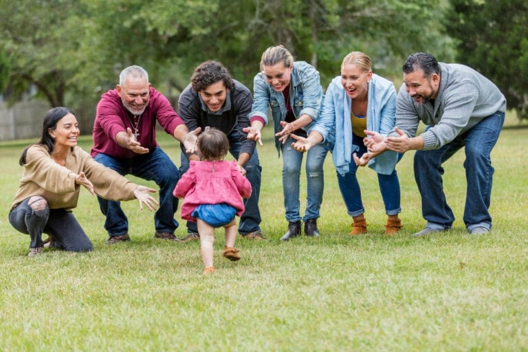 A multi-generation Hispanic family standing in the park together. A 16 month old baby girl is the center of attention. Her back is to the camera as she walks toward her family, all smiling with their arms open, reaching for her.