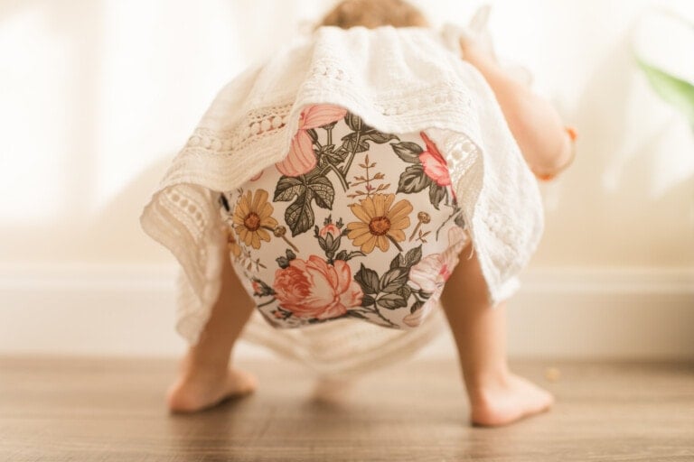 Toddler Crouched Down, Looking for Something While Wearing a Vintage Floral Modern Reusable Cloth Diaper and a White Shirt with a Neutral Wood Background in Bright Natural Light
