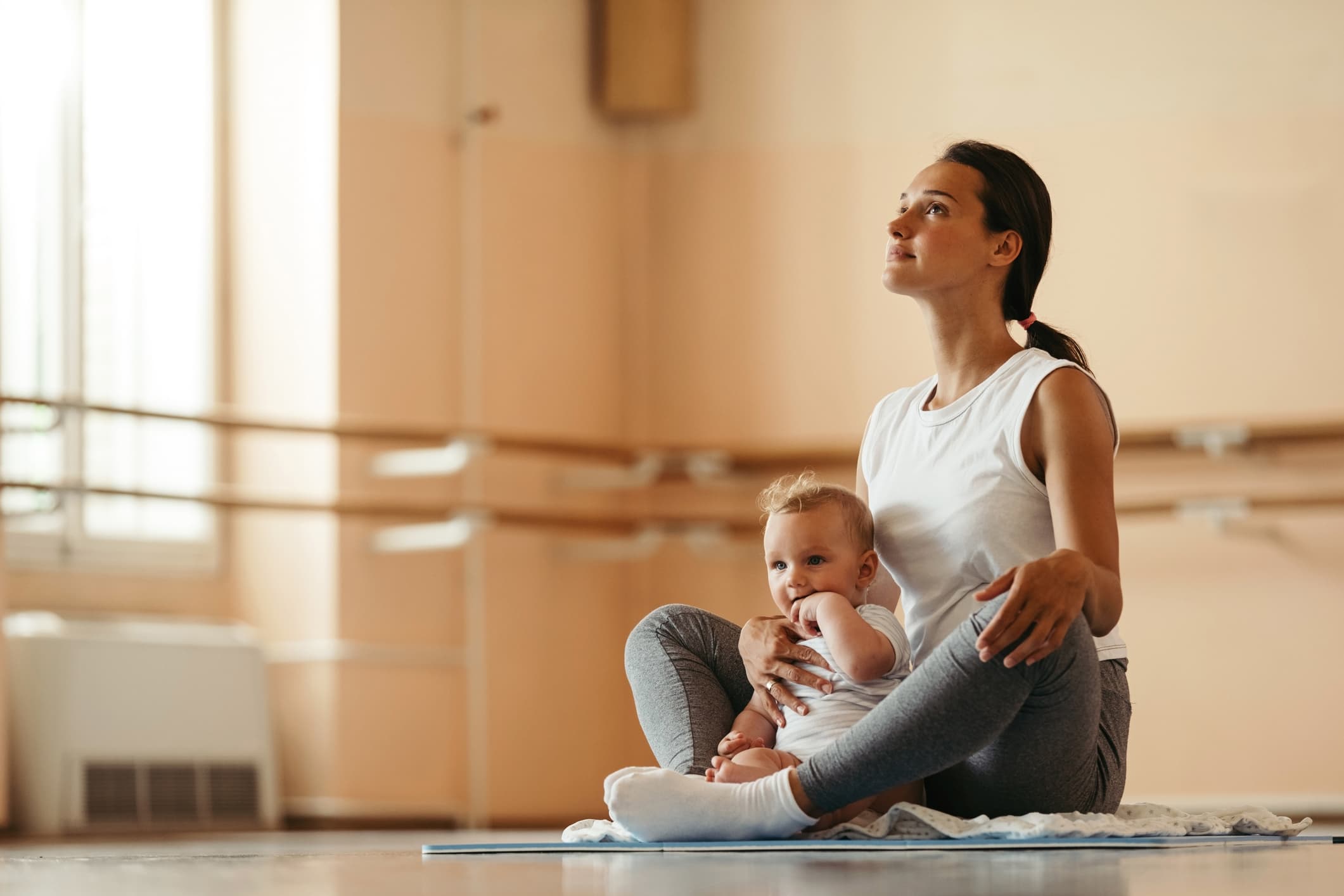Young mother sitting in lotus position while meditating with her baby in health club.