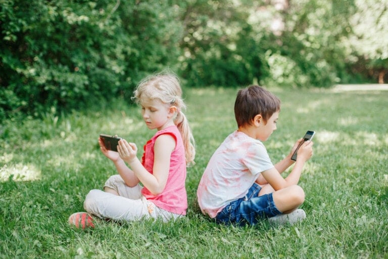 Boy and girl play games on smartphones outdoor. Kids digital gadget screen addiction. Children friends playing online sitting on grass in park.