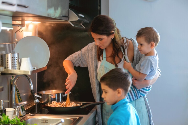 A woman is cooking on a stove while holding a young child on her hip, managing the precious hours in the day. Another child stands nearby watching. The kitchen is warmly lit, with various utensils and cookware scattered around the stove area. She appears to be stirring or frying food in a pan.