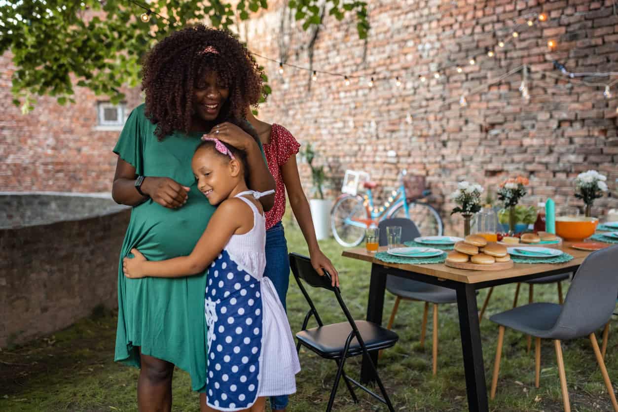 A woman in a green dress stands outside, her pregnant belly being hugged by a young girl in a polka dot dress. They are under a tree, near a table set with plates, glasses, and food, including a stack of buns. A bicycle is visible in the background as they celebrate the new mom.