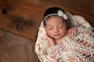 A three week old newborn baby girl sleeping in a little, wooden bowl. She is wearing a cream colored bow headband and swaddled with a decorative wrap. Shot in the studio on a wood background.