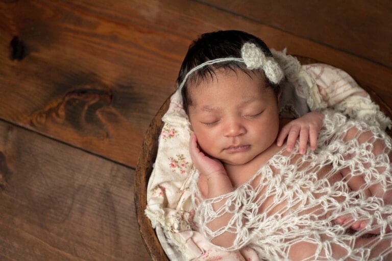 A sleeping baby with short black hair rests in a round wooden bowl, wrapped in a white knit blanket. The baby has a delicate headband with a small flower decoration and is lying on floral patterned fabric. The background is a wooden surface, evoking the charm of biblical girl names like Ruth or Esther.
