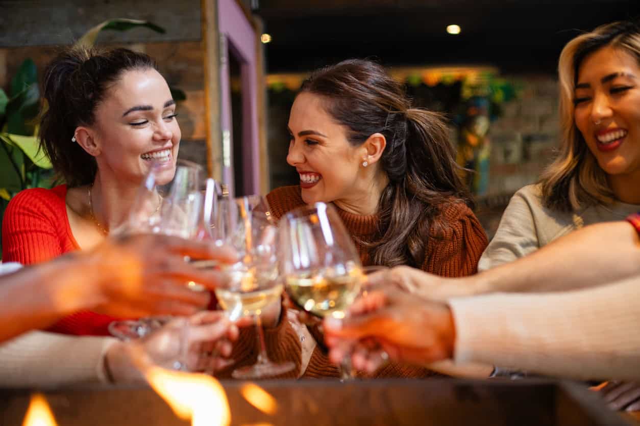A group of people are seated around a table, smiling and raising glasses in a toast. The setting appears to be a cozy indoor space with warm lighting. Some are holding glasses of white wine. The mood is festive and cheerful as they celebrate the new mom.