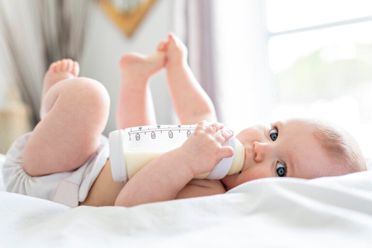 A Pretty baby girl drinks milk from bottle lying on bed. Child wearing a diaper in nursery room.