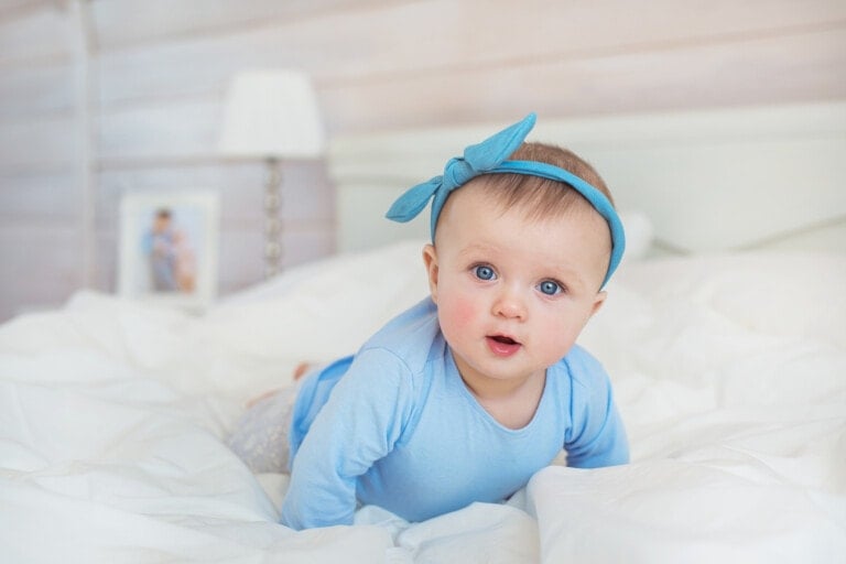 A baby wearing a blue outfit and a matching headband with a bow is lying on their belly on a white bed. The baby, possibly with one of those charming Russian girl names like Anastasia or Elena, has blue eyes and a curious expression. In the background, there is a blurred frame with a photo and a lamp.