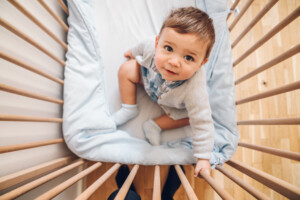 A toddler with short brown hair is sitting in a wooden crib, looking up at the camera. The crib has light blue bedding, and the baby is dressed in a light blue and white outfit. Despite signs of 9-month sleep regression, the crib remains cozy and calm on its wooden floor.