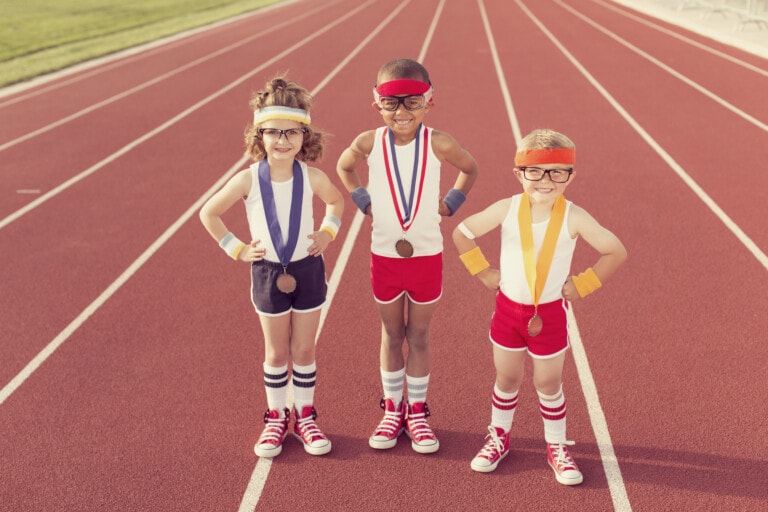 Three children in athletic clothing stand on a running track, beaming with pride. Each child wears a medal around their neck, colorful wristbands, headbands, glasses, and red sneakers. They are smiling and posing with their hands on their hips after an exhilarating competition. The track stretches out behind them.
