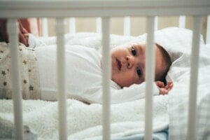 A baby wearing a white long-sleeved onesie and patterned pants lies on a soft white blanket in a crib. The baby, who seems to be waking up early, looks up with wide eyes, and the crib bars are visible in the foreground of the image.