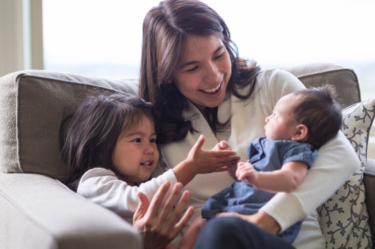 A woman is sitting on a couch holding a baby, part of her daily routine for newborn and toddler, and looking at a smiling young girl beside her. The girl reaches out her hands toward the baby while they both share a warm moment in the cozy living room.