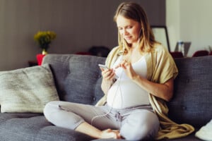 Positive expectant mother wearing earphones connected with cellphone, listening to music on smartphone. Pregnant young woman spending leisure time at home.