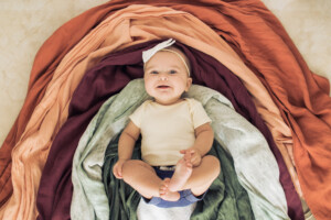 A 5-month-old rainbow baby girl wearing blue shorts, a light yellow shirt and a white bow on top of different color fabric making a warm-colored Autumn rainbow.