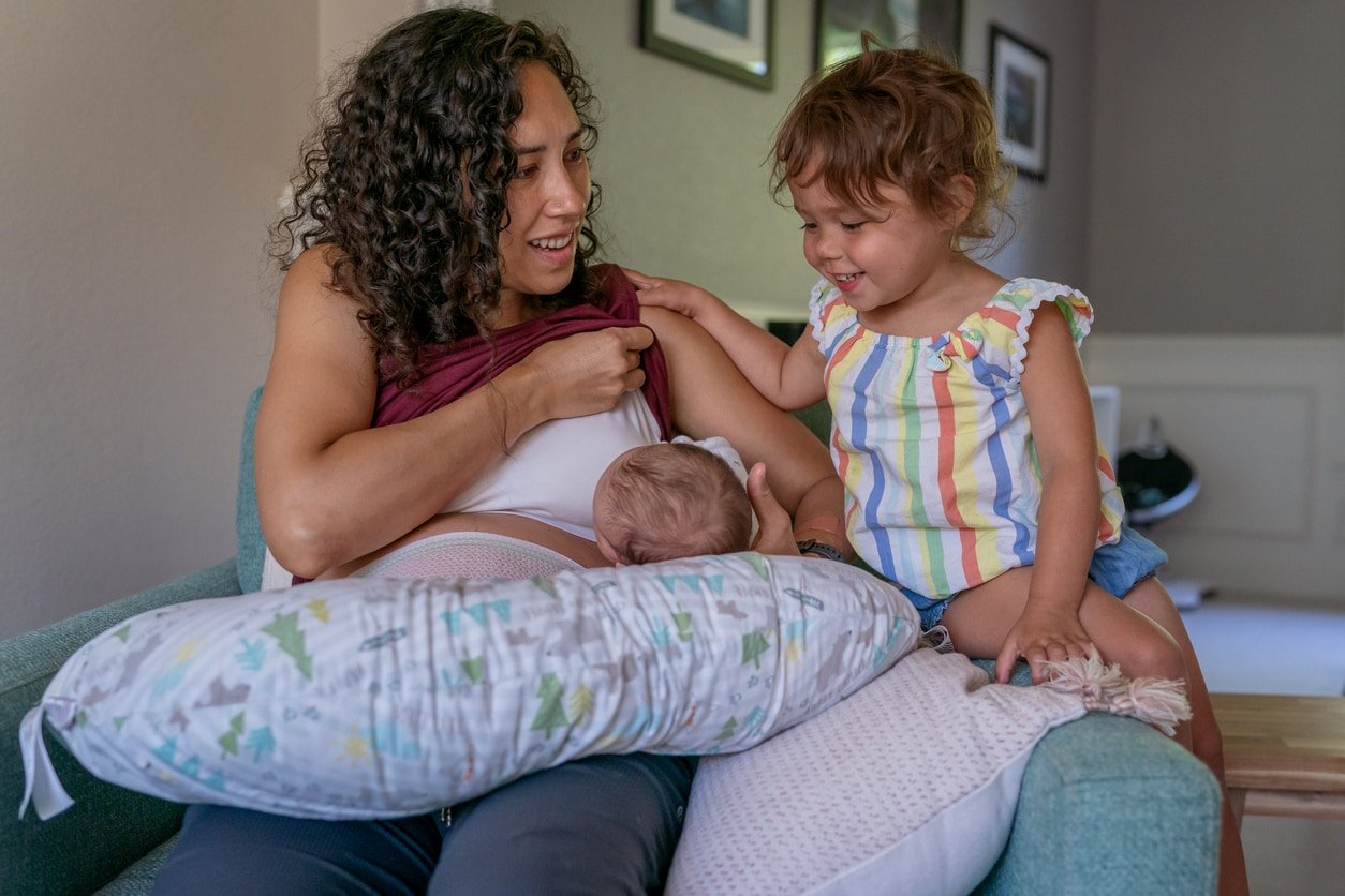 A multiracial woman sits on a chair at home and breastfeeds her baby while talking with her cute toddler daughter who is watching with curiosity.