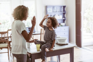 Happy little boy giving high-five to mother at home. Young woman is standing while son sitting on table. They are wearing casuals.
