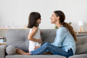 Attractive woman and little girl sitting on comfortable couch at home. Young mother talking with small adorable daughter storytelling.