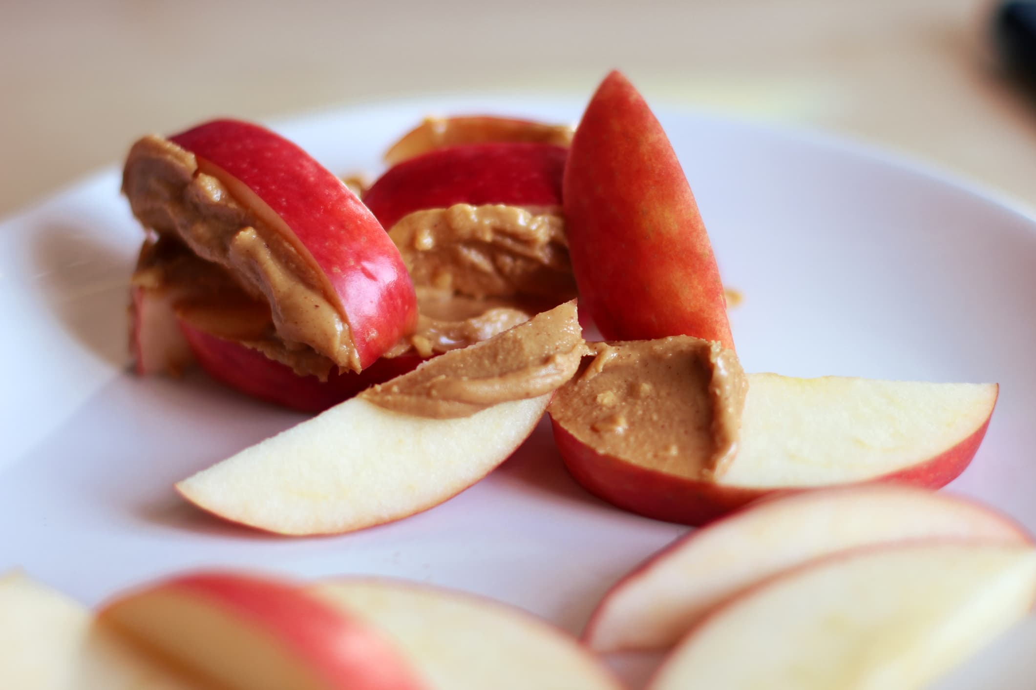 Sliced red apple with crunchy peanut butter spread on top. Served on a white plate with a pale background.