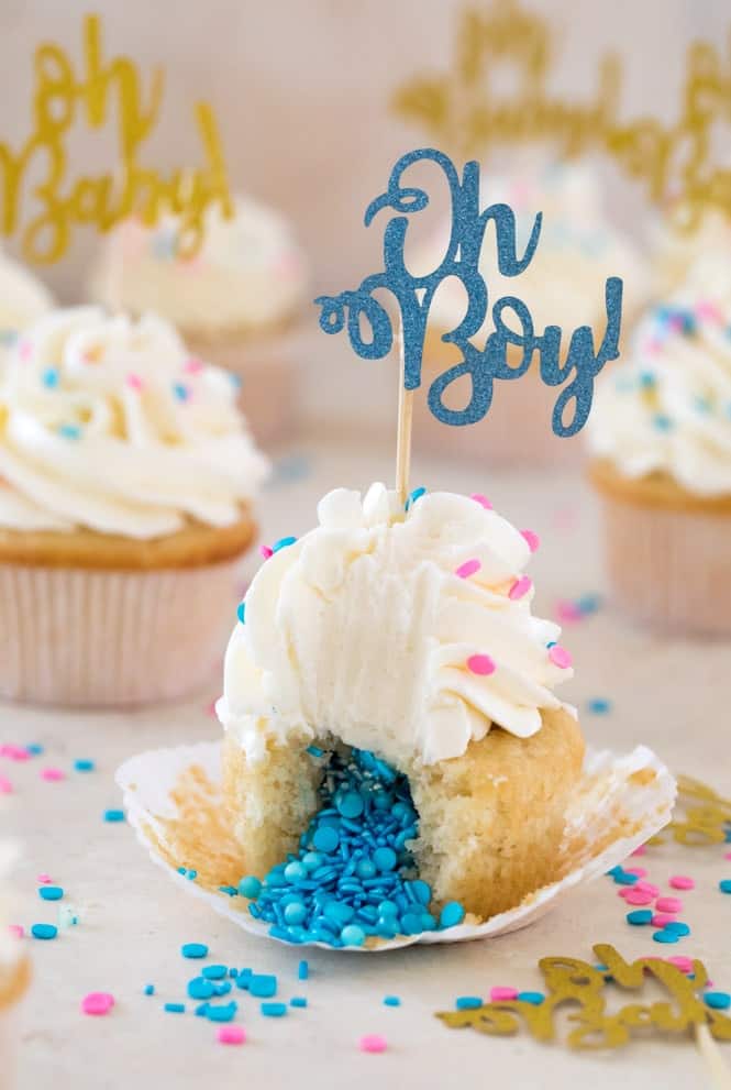 Vanilla cupcakes with white frosting and a blue Oh Baby! topper make for delightful gender reveal ideas. One cupcake, partially unwrapped, unveils blue sprinkles inside. Pink and blue sprinkles are scattered across the table, adding to the festive surprise.