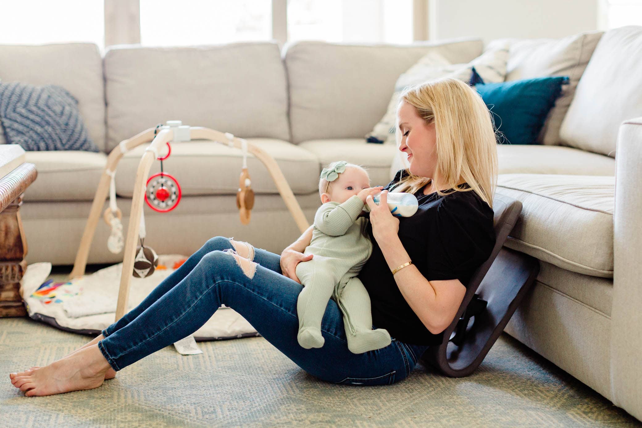 Woman sitting on the floor feeding her daughter a bottle while using the Ready Rocker against the sofa.